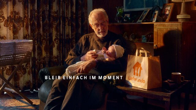 Grandfather holding a baby in an armchair, a Lieferando paper bag on the table beside him