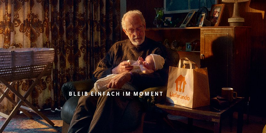 Grandfather holding a baby in an armchair, a Lieferando paper bag on the table beside him