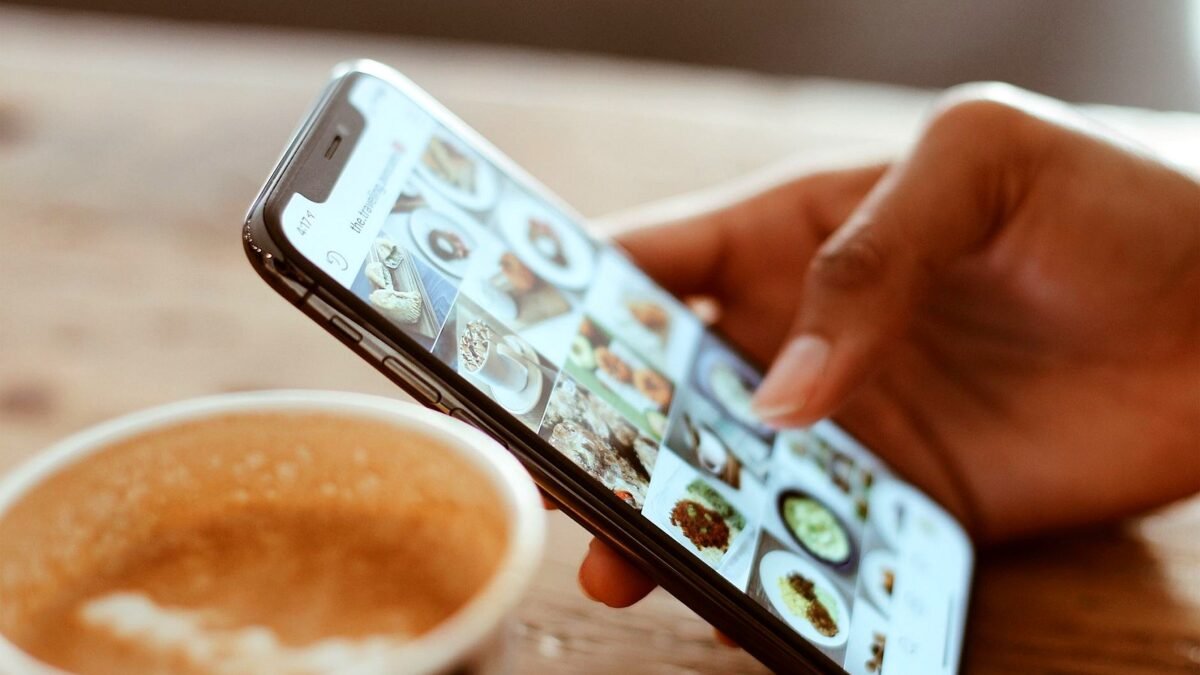A hand scrolls through photos in a social media app on a smartphone, next to a cup of coffee
