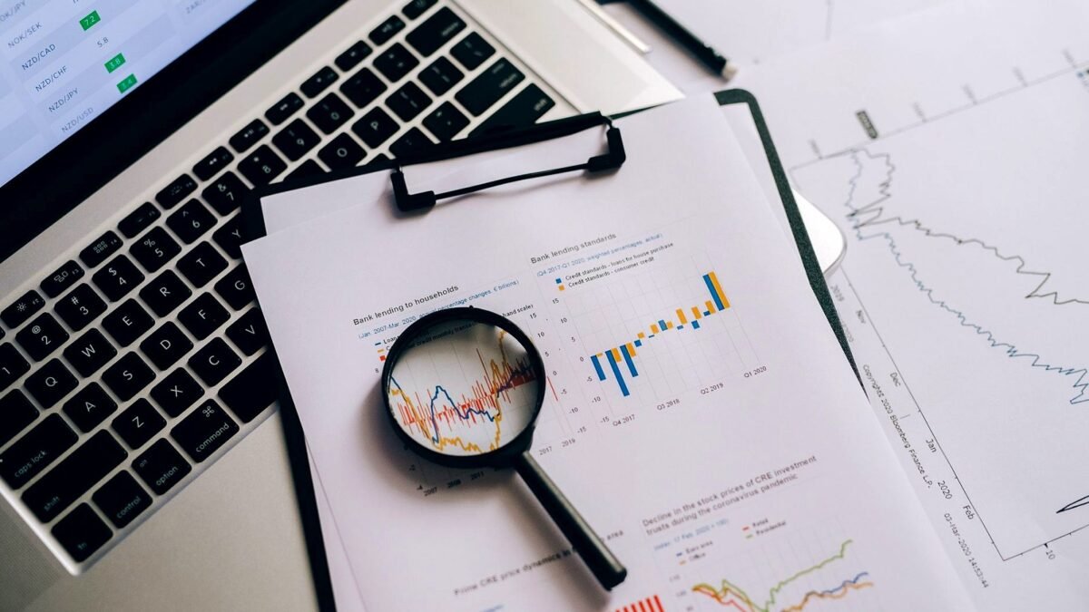 Laptop, financial charts and a magnifying glass on a desk seen from above