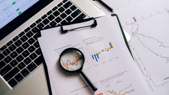 Laptop, financial charts and a magnifying glass on a desk seen from above