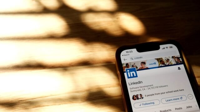 A smartphone on a wooden table shows a LinkedIn profile page