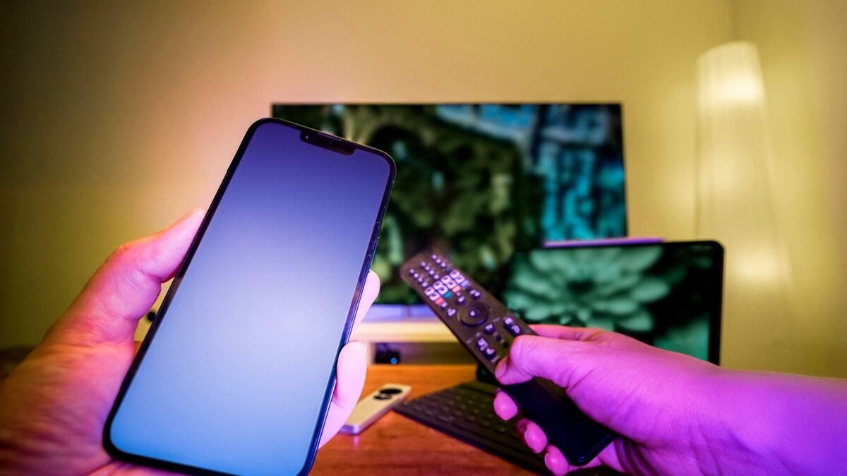 Hands holding a smartphone and a TV remote in a living room