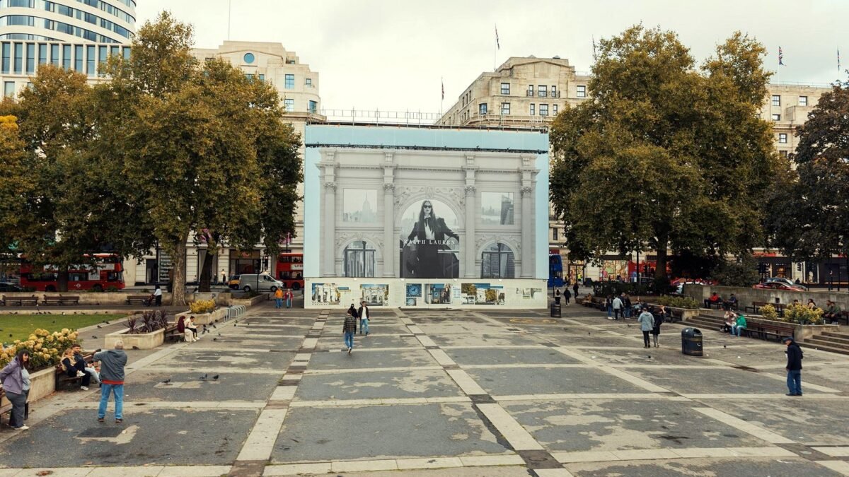 Red London buses pass a large advertising screen on a city square