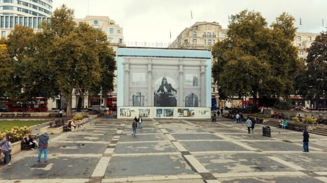 Red London buses pass a large advertising screen on a city square