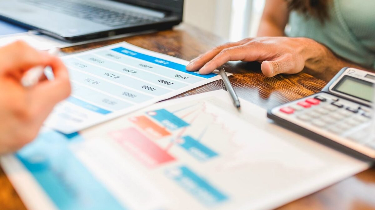 Hands at a desk point at printed financial charts next to a calculator and pen