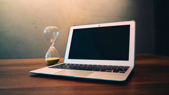 Laptop on a wooden desk next to an hourglass with running sand